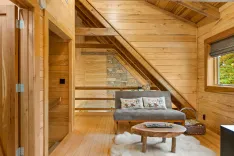 Cozy wooden cabin interior with a gray sofa, rustic wooden coffee table on a fluffy white rug, and stairs leading to an upper level.