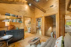 Cozy wooden cabin interior with an open floor plan, including a living area with a bookshelf and a sofa, a visible bedroom, and warm lighting.