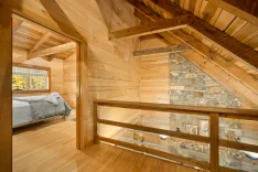 Interior of a cozy wooden cabin with exposed beams, featuring a bedroom with a bed visible through a glass balustrade and a stone wall accent.
