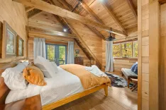 Cozy rustic cabin bedroom with a vaulted ceiling, exposed wooden beams, hardwood floors, and a large bed with white linens and an ochre blanket.