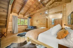 Cozy wooden cabin bedroom interior with a large bed, cowhide rug on the floor, exposed beams on the ceiling, a seating area with a grey sofa, and natural light coming through the windows.