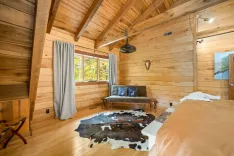 Rustic cabin-style bedroom interior with wooden walls and ceiling, furnished with a brown leather bed, a gray sofa with blue pillows, a cowhide rug on the floor, and a ceiling fan above. Natural light streams in from windows overlooking greenery.
