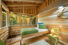 Cozy wooden cabin interior with a hanging bed, floor-to-ceiling windows showcasing autumn foliage, a green chair with lamp, and rustic decor.