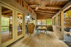 Cozy screened porch with wooden floors, walls, and ceiling, rustic fan above, glass doors, furnished with a table and chairs, overlooking autumn trees.