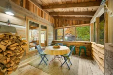 Cozy enclosed porch with a rustic wood interior, a round table with chairs, and a stack of firewood, overlooking autumn trees.