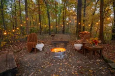 Cozy autumn outdoor setting with a fire pit, surrounded by chairs with blankets, string lights amongst trees, and colorful fall foliage.