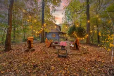 Cozy autumn backyard scene with string lights, picnic table, and a wooden cabin surrounded by trees with fall foliage.
