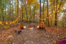 Cozy autumn outdoor seating area with fire pit, string lights, wooden benches, and seasonal decorations surrounded by fall foliage.