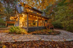 A cozy wooden cabin with lit interior and string lights at twilight, surrounded by autumn foliage.