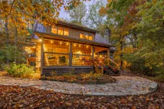 Cozy wooden cabin with lit interior surrounded by autumn foliage during twilight.
