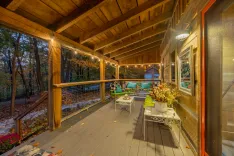 Cozy wooden porch decorated with string lights, outdoor furniture, and plants during evening time, surrounded by autumnal trees.