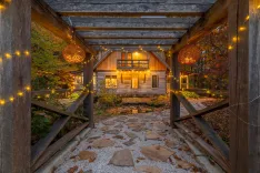 A cozy wooden cabin at dusk, warmly lit from the inside, with a stone path leading to the entrance framed by a rustic wooden pergola adorned with string lights and hanging glass lamps.