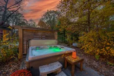 A cozy outdoor hot tub with illuminated green waters set against a backdrop of autumn foliage and a vibrant sunset sky.