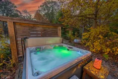 A hot tub with green lighting set against an autumnal forest backdrop during sunset, with a decorative lantern casting patterns on a wooden surface.