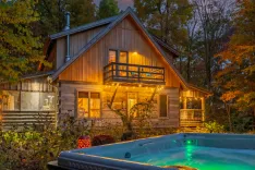 Cozy wooden cabin with illuminated interior and string lights at dusk, surrounded by autumn foliage and a hot tub in the foreground.