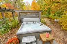 Outdoor hot tub on a wooden deck surrounded by autumn foliage with a pink speaker on a side table.