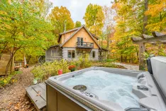 A cozy wooden cabin with a balcony surrounded by trees with autumn foliage and a steaming hot tub in the foreground.