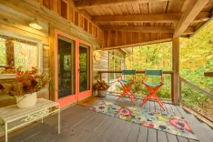 Cozy wooden cabin porch with two red chairs, decorative autumn-themed pot on bench, and 'Hello' doormat, surrounded by fall foliage.