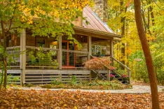 Cozy wooden cabin with a front porch, surrounded by autumn foliage and fallen leaves, with a stone chimney and red roof accents.