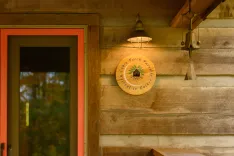 A warm outdoor light fixture illuminates a wooden plaque with the inscription "Cabin Porch Porch Package. Retreat to Nature" on a rustic cabin wall near an orange door.