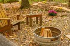 Wooden Adirondack chair and side table next to a metal fire pit with stacked wood, surrounded by fallen autumn leaves.