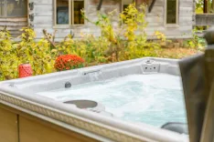 Outdoor hot tub filled with bubbling water near a garden with a decorative pink can on the edge, set against a rustic house backdrop.