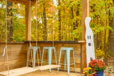 A cozy outdoor porch decorated for autumn with a wooden swing, teal barstools, a decorative ghost figure, and vibrant fall foliage in the background.