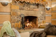 Cozy cabin interior with a lit fireplace, stone surround, antler decor, and fireplace tools set to the side.
