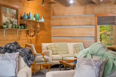 Cozy cabin living room interior with rustic wooden walls, comfortable sofas with plaid cushions, and decorative pottery on shelves.
