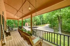 Spacious wooden porch with rocking chairs and a swing overlooking a forested area with a gravel driveway.