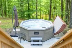 An outdoor hot tub with bubbling water on a patio surrounded by trees, with a folded lounge chair and a closed patio umbrella nearby.