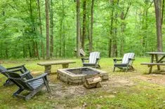 A peaceful outdoor camping area with a fire pit, Adirondack chairs, and a picnic table surrounded by lush green forest.