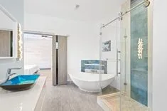 Modern bathroom interior with a unique blue glass vessel sink, white freestanding tub, glass-enclosed shower area, and artistic wall details.