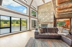 Spacious living room with high ceilings, large windows overlooking a lake, exposed wooden beams, stone fireplace, and a leather couch.