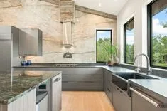 Modern kitchen interior with granite countertops, stainless steel appliances, and large windows overlooking greenery.