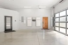 A clean and modern garage interior with a Tesla home battery setup on the wall, a wooden door, and a glass-panel garage door.