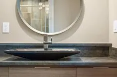 Modern bathroom interior with a round mirror and a black vessel sink on a gray countertop with wall-mounted faucet.