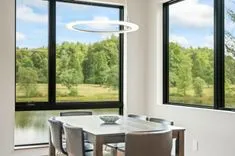 Modern dining room with large windows overlooking a green landscape, featuring a round pendant light above a wooden table with chairs.