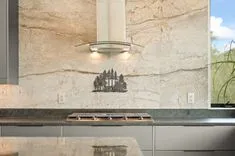 A modern kitchen backsplash with beige marble tiles, featuring a stainless steel range hood, decorative metal tree silhouette, and a gray countertop with a built-in gas cooktop.