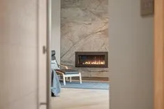 View from a partially open door towards a modern living room with a comfortable chair and lit gas fireplace set in a stone wall.