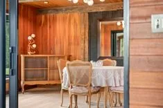 Elegant dining room interior with a wooden table set with delicate lace cloth, vintage chairs, and wooden cabinet against a rustic wooden wall.