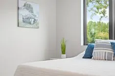 Tranquil bedroom corner featuring a neatly made bed with blue and striped pillows, a potted plant on a bedside table, and a canvas of a floral truck hanging on the wall, with a window providing a view of greenery outside.