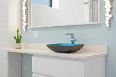 Modern bathroom with a blue vessel sink, chrome faucet, and a small cactus on the counter, with lit vanity mirror lights in the background.