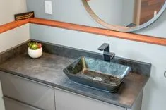 Modern bathroom with a rectangular vessel sink, dark matte faucet, gray countertop, wooden shelf with motivational sign, round mirror, and a small potted plant.