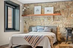 Cozy bedroom interior with stone wall, wooden shelf with framed pictures, bed with pillows and striped throw, and vintage stove on the side.