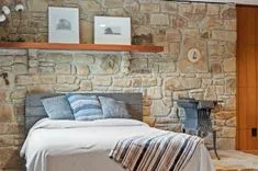 Cozy bedroom interior with a stone wall, wooden shelf with framed art, a cast-iron stove, and a bed with grey and blue pillows and striped blanket.