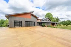 Modern house with a large concrete driveway, attached garage with two black doors, and a combination of wood and metal exterior under a blue sky with clouds.