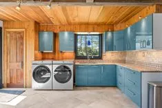 Bright and modern kitchen with blue cabinets, stainless steel appliances, and a wooden ceiling. A pair of front-loading washing machines indicates a combined laundry space.