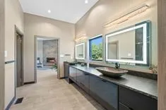 Contemporary bathroom interior with double vanity, vessel sink, backlit mirrors, and a view into a bedroom with a fireplace through an open door.