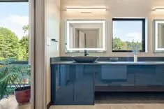 Contemporary bathroom interior with a view of a lush landscape through the window, featuring a dark vanity with a vessel sink, illuminated by overhead sconce lighting, and framed mirror.